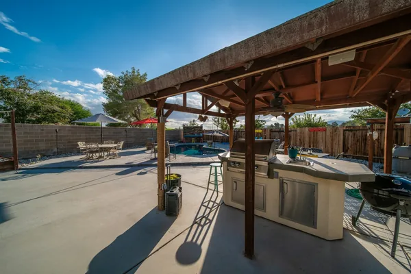 Large backyard concrete patio with an outdoor kitchen under a wood cover, a pool and a block wall behind