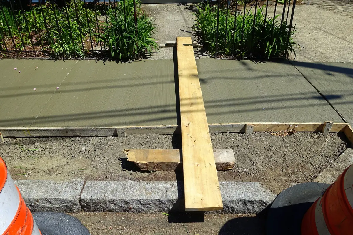 Freshly poured sidewalk panel between wood forms, with a board laid across it